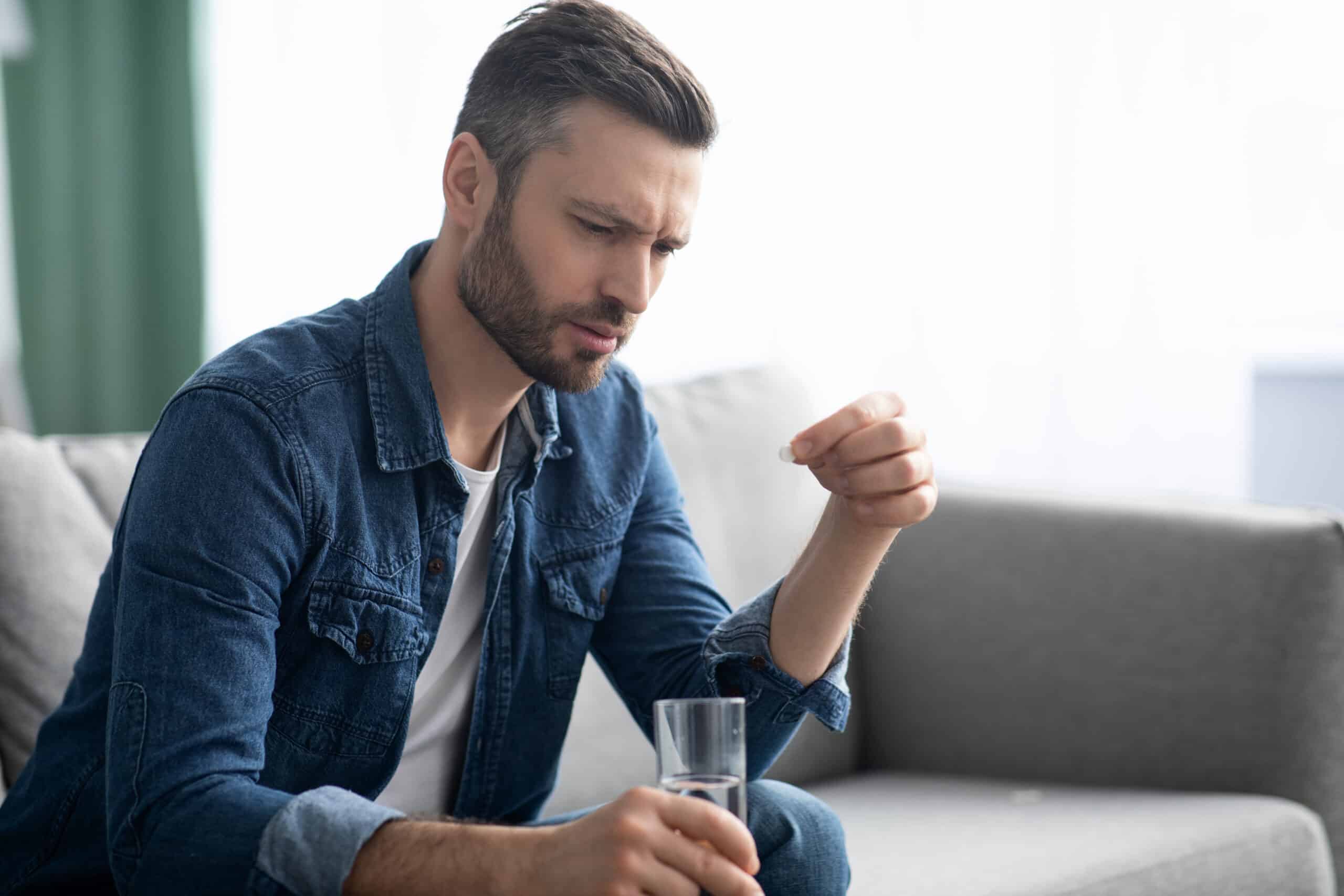 Upset bearded man with glass of water taking medicine